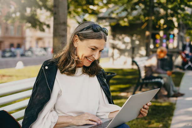 人、年龄、生活方式、技术和通讯。户外照片中，美丽的退休女性穿着时尚的衣服，坐在长凳上，膝盖上放着触摸板电脑，看着有趣的视频，笑着图片下载