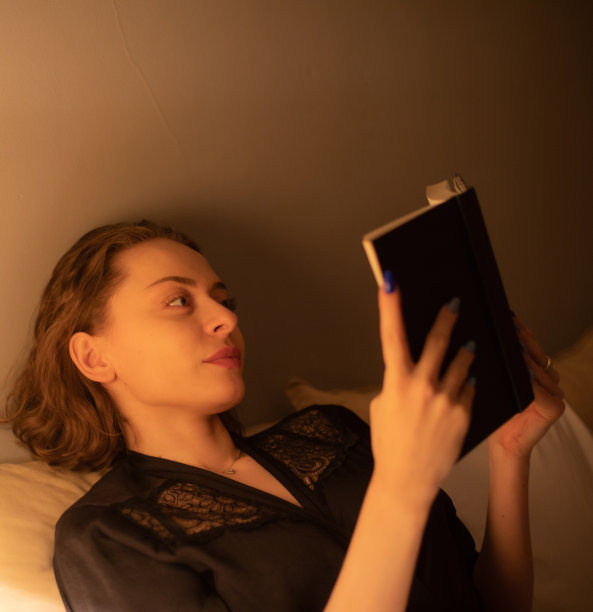 年轻美丽的女人坐在她的卧室里读一本书，她睡觉前，放松在她舒适的床上。图片下载