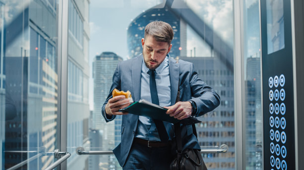商务经理上班迟到，乘坐现代办公大楼的玻璃电梯。帅哥吃着美味的外卖汉堡乘电梯赶去公司开会。图片下载