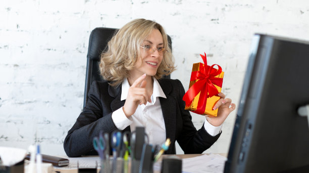 在工作场所给自己一个惊喜。快乐的成年女子穿着正式的衣服，手里拿着一个红色的礼品盒放在办公室的办公桌上，对着电脑屏幕，选择性的专注。图片下载