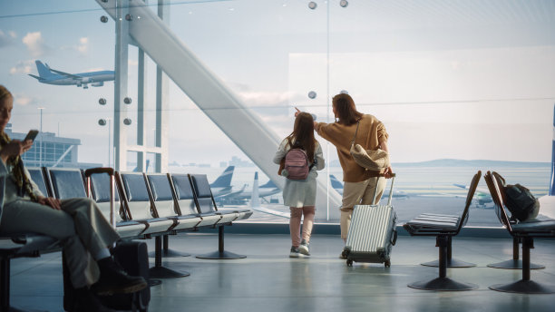 机场候机楼:美丽的母亲和可爱的小女儿在等待他们的假期航班，看着窗外到达和离开的飞机。航空枢纽登机休息室的年轻家庭图片下载