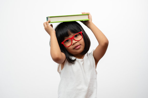 有趣和快乐的亚洲学龄前小女孩，戴着红色的眼镜，头上拿着一本绿色的书，在白色孤立的背景。学龄儿童的概念和小学、幼儿园、家庭学校的教育图片下载
