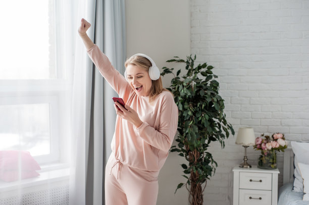 身穿家居服、戴着白色耳机、手里拿着智能手机的金发中年女性举起了手，表示了强烈的喜悦。胜利或好运的消息。图片下载