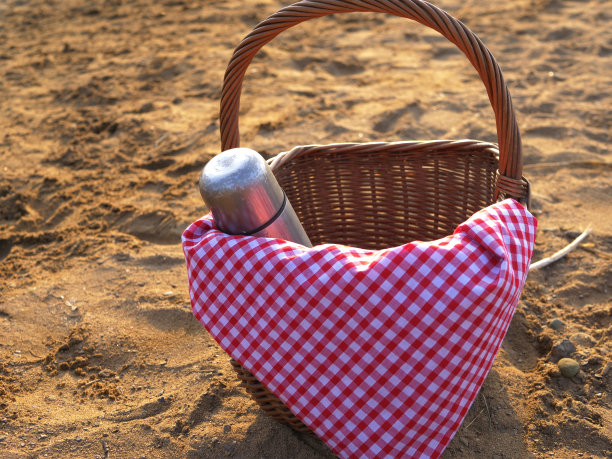野餐篮和保温瓶放在沙滩上的红色格子布上图片下载