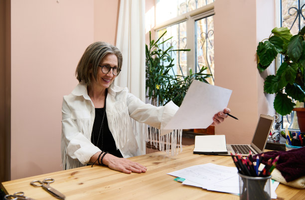 一位富有灵感的成熟商业女性，女性时装设计师坐在她的设计工作室的桌子旁，微笑着看着画的草图，露出欢快的牙齿图片下载