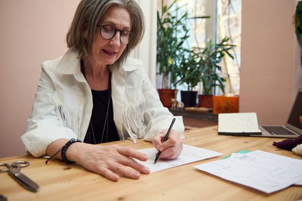 优雅成熟的女性时尚设计师在设计工作室绘制新服装系列的草图，女性裁缝创作线条，在木制的个人书桌上用铅笔和水彩笔作画图片下载