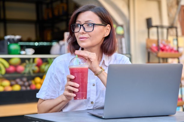 一位中年妇女在户外果汁吧的桌上用笔记本电脑喝新鲜果汁图片下载