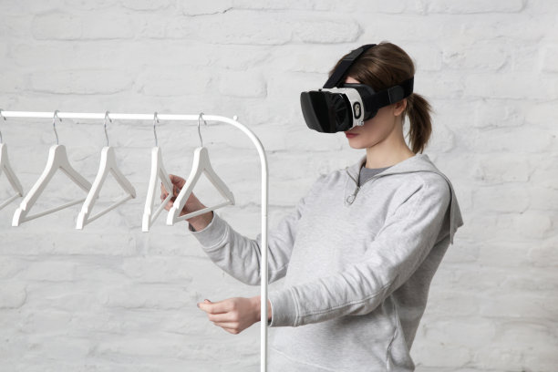 戴着VR眼镜从衣架上挑选服装的女人的肖像。虚拟现实体验与未来技术概念。图片下载
