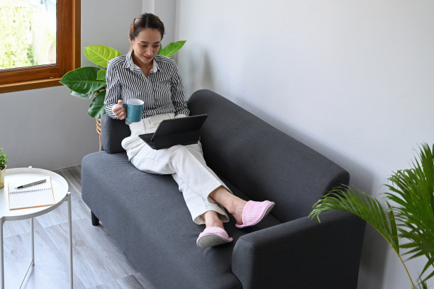 休闲的年轻女子坐在沙发上使用平板电脑，在家里花时间在社交媒体上。图片下载