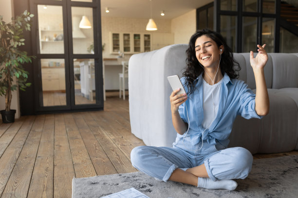 快乐的年轻女子戴着耳机用手机听音乐或有声读物，坐在家里的地板上，自由自在图片下载