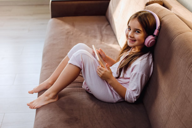 一个戴着粉色耳机的漂亮小女孩坐在沙发上，看着一台数码平板电脑图片下载