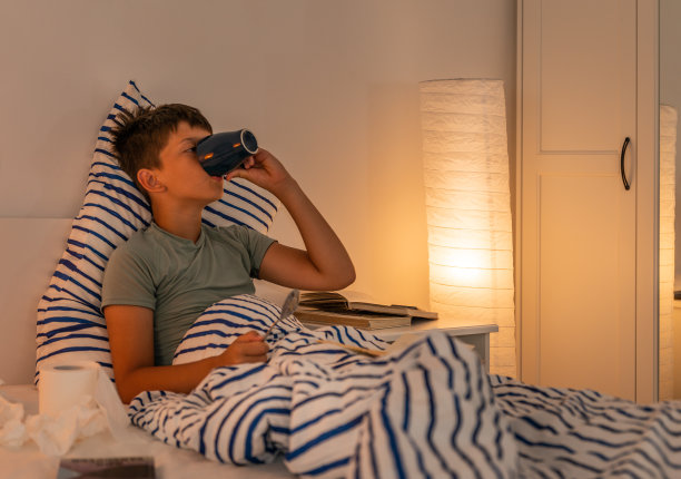 一个生病的少年躺在床上，身边围着手帕和一卷厕纸，伤心地擦着鼻子，读着一本书。图片下载