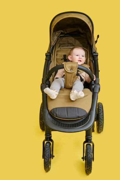一个快乐的孩子坐在画室黄色背景的婴儿车里。微笑的蹒跚学步的小男孩，在一个变压器婴儿车运送儿童，8个月大图片下载