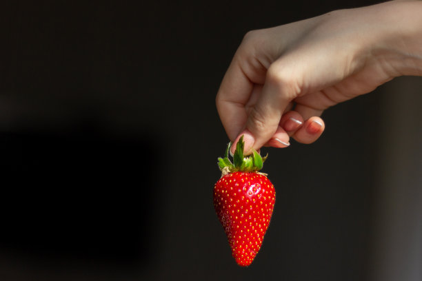 一个大的成熟多汁的红色草莓，在黑色的背景下，一个女性的手拿着它的尾巴。夏日浆果。健康的食物图片下载