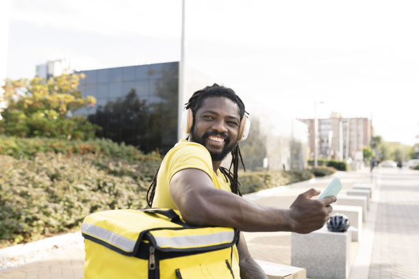 穿着黄色制服的年轻西班牙裔送货员带着耳机微笑地听着音乐，从纸盒里拿出一个纸盒。外观。图片下载