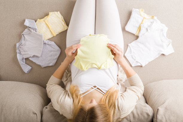 年轻的孕妇大肚子坐在客厅的米色沙发上，为未来的新生儿整理衣服。在怀孕期间准备东西。孩子的期望。自顶向下的观点。图片下载