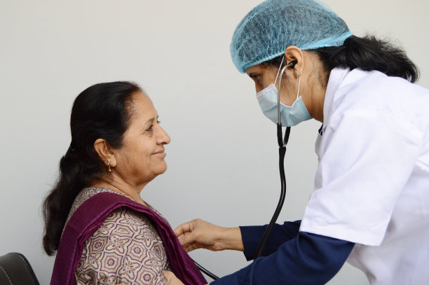 戴着外科手术口罩、帽子、白大褂、脖子上挂着听诊器为病人做体检的妇女或女医生，背景为灰色，有文字复制空间图片下载