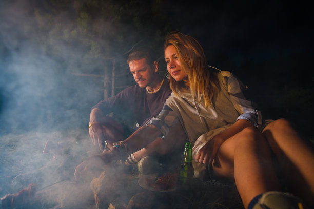 年轻的夫妇在篝火上烤香肠，在黄昏的森林小山上喝啤酒图片下载