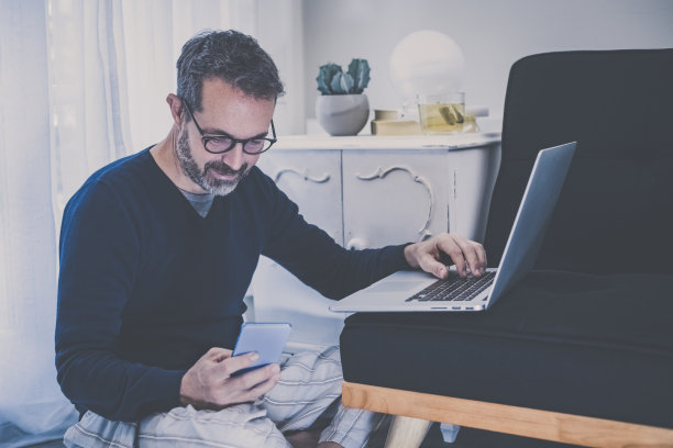 一名男子坐在家里的地上用笔记本电脑工作。男性在家工作，有远程办公室。男孩在客厅使用网络设备。家庭作业、家庭教育生活方式新传播理念图片下载