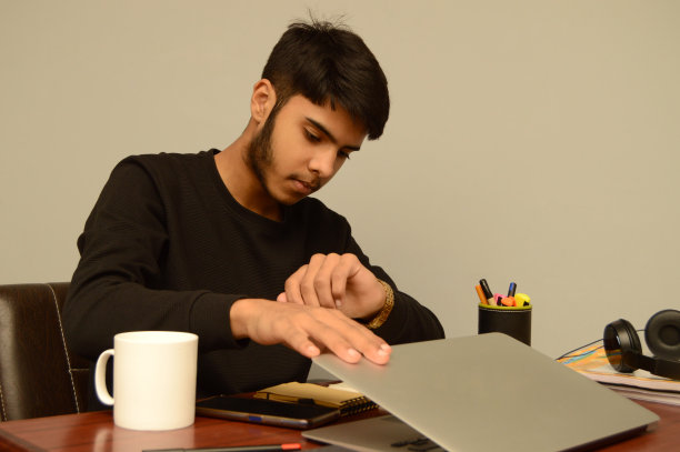 一名黑发胡须的年轻男性一边看手表一边戴着耳机合上办公桌上的笔记本电脑，灰色背景下他的办公桌上放着一台笔记本电脑——关机或关机时间图片下载