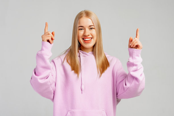 快乐时尚的金发女孩在时髦的连帽衫指着手指和微笑，邀请注册，显示公司的标志，站在灰色的背景，提供信息图片下载