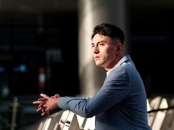 英俊的中国年轻人的肖像在浅蓝色的西装和白色的汗衫看着远离现代城市建筑背景在阳光明媚的日子，侧面的观点自信的商人。图片下载