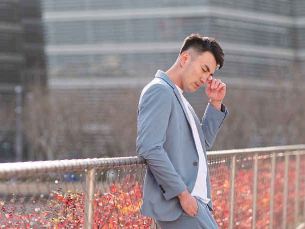 在阳光明媚的日子里，穿着浅蓝色西装和白色汗衫的英俊中国年轻人用手指揉着眼睛，以现代城市建筑为背景，侧面是疲惫的商人。图片下载