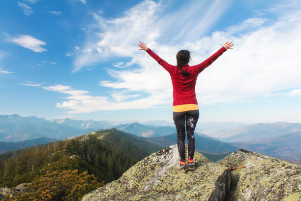 山顶上的年轻女孩在蓝色的天空背景下举起了手。那个女人爬到了顶峰，享受着她的成功。后视图图片下载