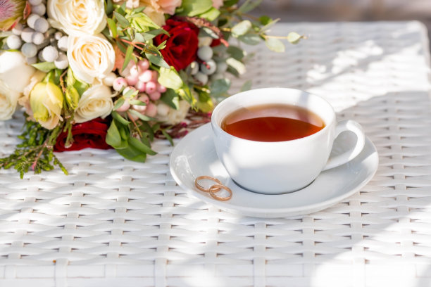 一杯茶，玫瑰花瓣，金婚戒和一束漂亮的花束放在白色的桌子上。婚礼庆典的概念。新娘早上好。图片下载