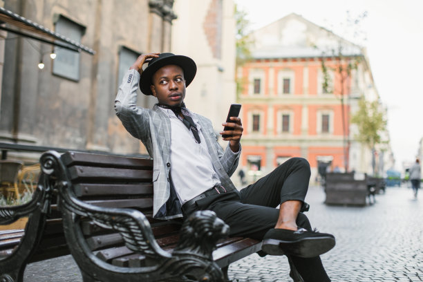 穿着西装、戴着帽子的非洲男子坐在长椅上，手里拿着智能手机图片下载