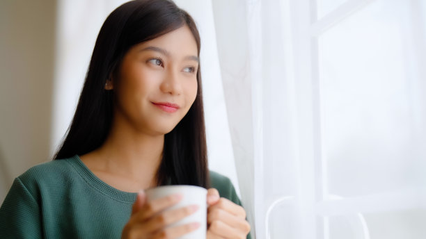 年轻的亚洲妇女站在窗边，拿着杯子在卧室里，她早上醒来后喝牛奶图片下载