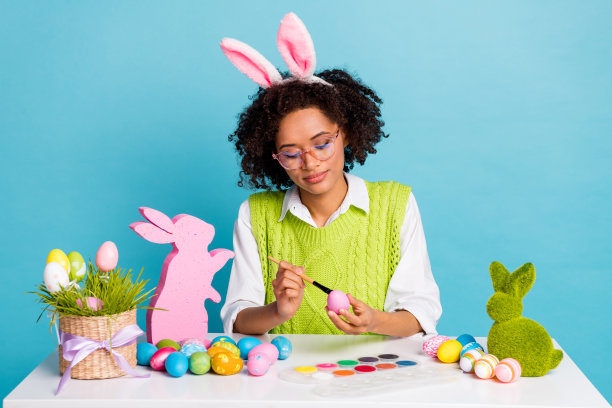 照片好的千禧一代深褐色的女士油漆鸡蛋穿绿色背心孤立在蓝色的背景色图片下载