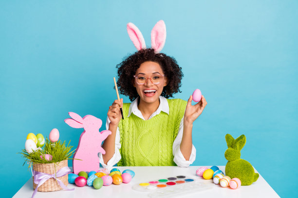 印象深刻的千禧一代深色皮肤的女士画鸡蛋穿绿色背心孤立在蓝色背景的照片图片下载