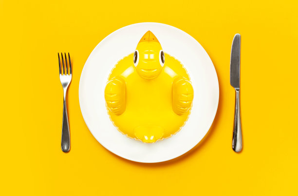 可充气的迷你黄色鸡或鸭子在白色的盘子，叉刀在黄色的背景。创意食品理念，烟草鸡。潮流充气游泳儿童玩具。布局设计图片下载