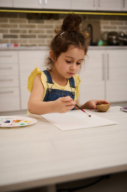 这是一个学龄前的可爱的欧洲小女孩，她坐在木桌前，用画笔在调色板上调出水彩画或水粉画图片下载