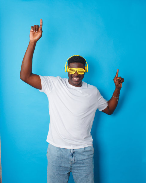 一个非裔美国人戴着黄色的耳机和时髦的眼镜，在一个孤立的蓝色背景下欢快地跳舞图片下载
