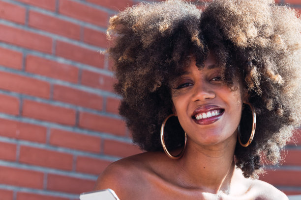 近距离的快乐非洲裔美国妇女看着相机，而持有智能手机户外。非洲裔女性有着完美的白色微笑，长长的非洲式发型和迷人的砖墙妆容图片下载