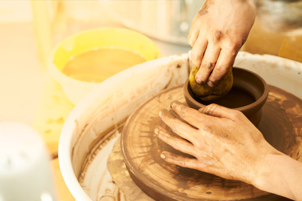 在作坊里，一个陶工在陶轮上用红粘土制作陶器。女人的手创造了一个罐子。女孩在陶罐特写雕塑。粘土特写。温暖的照片的气氛。穿围裙和格子衬衫的女孩。图片下载