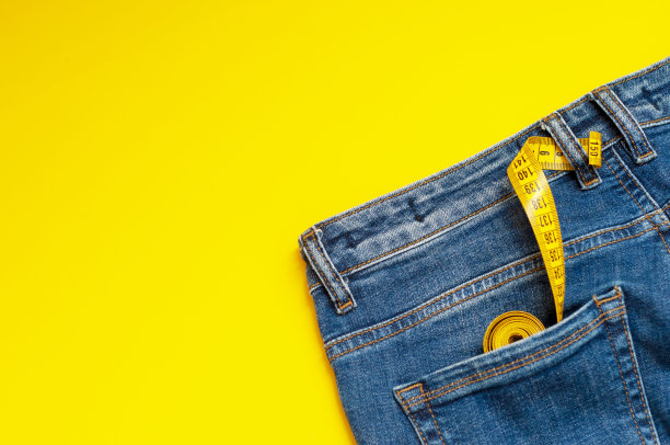 卷尺在蓝色牛仔裤的后口袋里，黄色背景，复印空间。概念饮食、体重控制、体重减轻、体重减轻、与体重减轻或增加有关的疾病、贪食症。图片下载