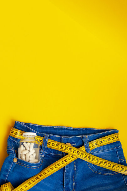 黄色的卷尺穿在蓝色牛仔裤上，而不是黄色背景上的腰带，裤子口袋里有一个装有药丸的玻璃瓶，一个复印空间，一张垂直的照片。图片下载