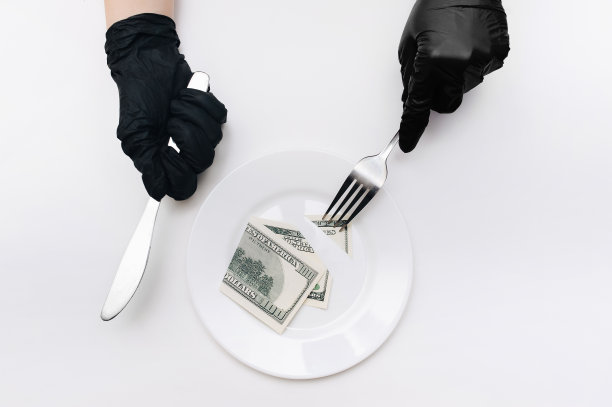 商务午餐的概念。戴着黑色手套的女性手拿着刀叉，从一张百元大钞上切下一块放在一个白色盘子上。设想可视化税收，贬值和利息。图片下载