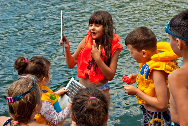 穿着泳装和充气救生衣的孩子们在水边玩耍。孩子们用笔记本电脑和其他小工具在大自然中放松。图片下载