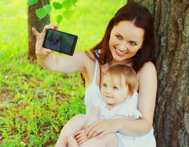 夏日公园的草地上，快乐微笑的母亲带着孩子用智能手机自拍图片下载