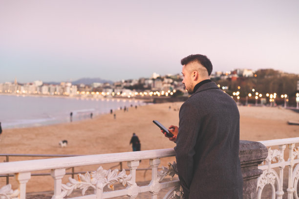 日落时分，在多诺斯塔-圣塞巴斯蒂安海滨散步，一名年轻的白人男子在使用智能手机。图片下载