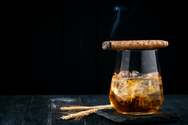 一杯加冰块的威士忌或干邑白兰地，上面放着一支冒烟的雪茄，黑色的木制背景图片下载