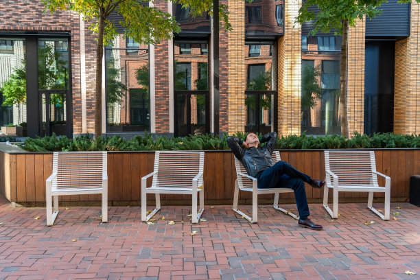 一名穿着长裤和黑色夹克的千禧一代男子正坐在四把椅子中的一把上图片下载