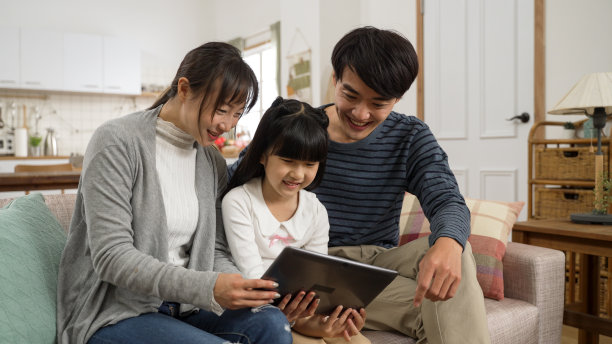 幸福的亚洲三口之家在家里的客厅里用平板电脑一起看在线视频。微笑的爸爸一边看一边评论图片下载