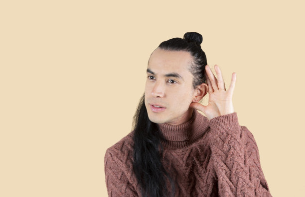英俊的年轻人穿着毛衣，在孤立的背景下微笑着，手放在耳朵上听谣言或八卦。耳聋的概念。图片下载