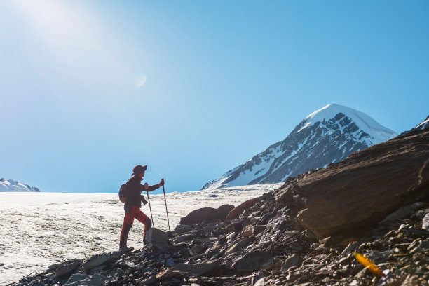 风景优美的高山景观，在阳光下，徒步旅行者的侧影与大冰川舌和雪山山峰。阳光明媚的日子里，背着背包的人在蓝天下的高山中行走。图片下载