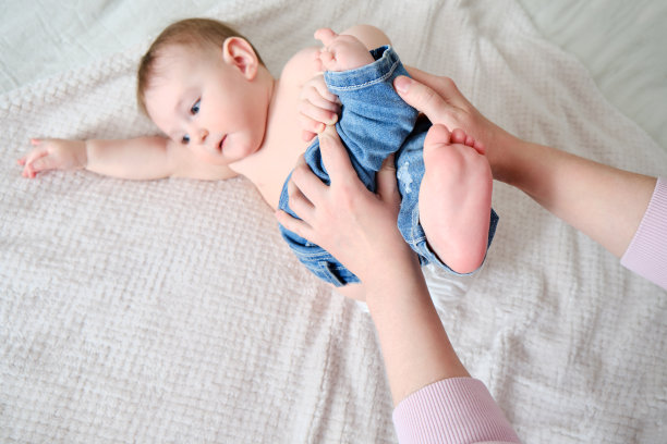 一位母亲为一个男婴穿上牛仔裤。妈妈在床上给一个快乐的孩子穿上衣服。图片下载